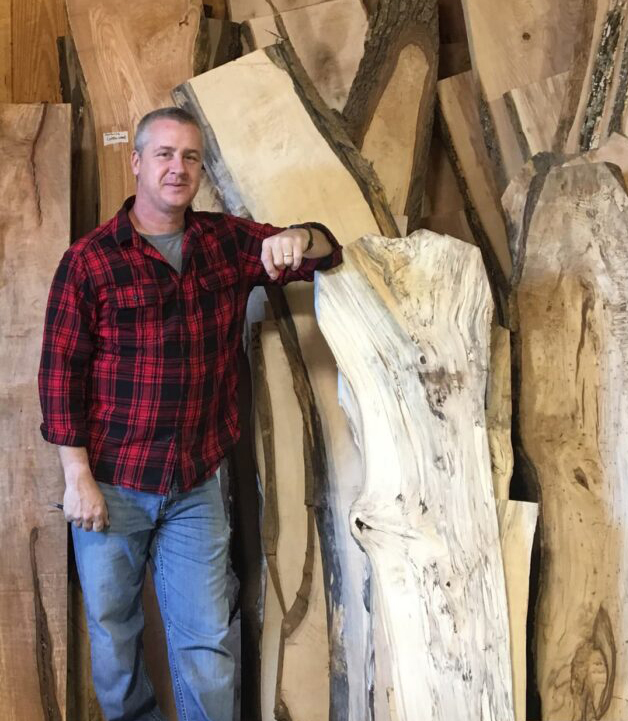 Aaron Moody, Owner, in a red and black flannel shirt standing in front of cut wood.